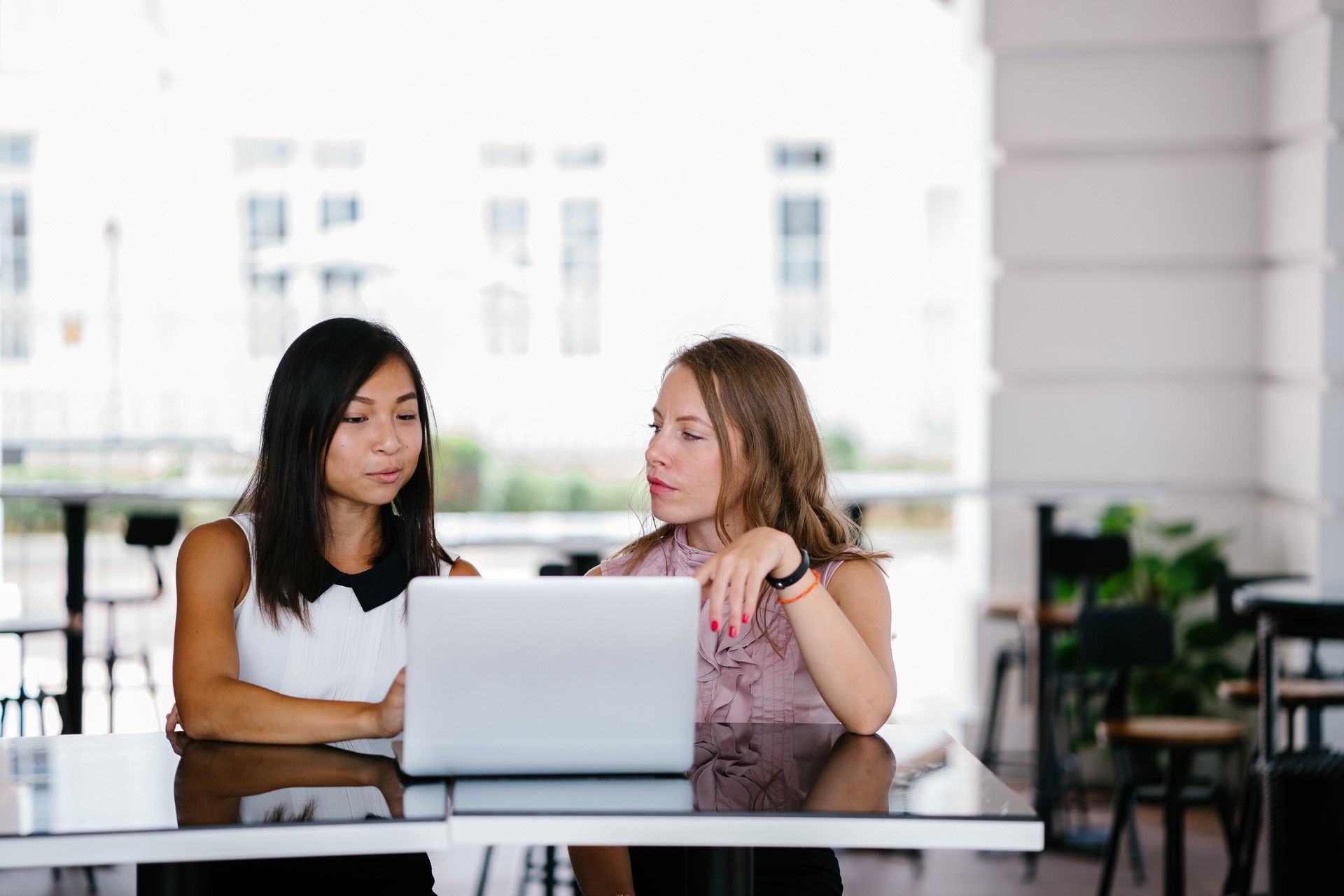 A diverse team of two coworkers (Chinese Asian woman and Caucasian white lady) working over a laptop. They are looking at the screen and discussing what is on the screen. 